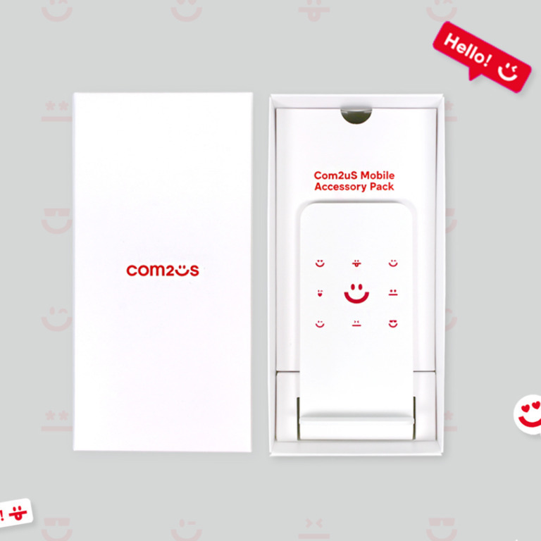 com2us 로고가 인쇄된 흰색 상자와 'Com2uS Mobile Accessory Pack'이라고 적힌 모바일 액세서리 팩이 나란히 놓여 있다. 액세서리 팩에는 여러 개의 빨간색 웃는 얼굴 아이콘이 그려져 있다.