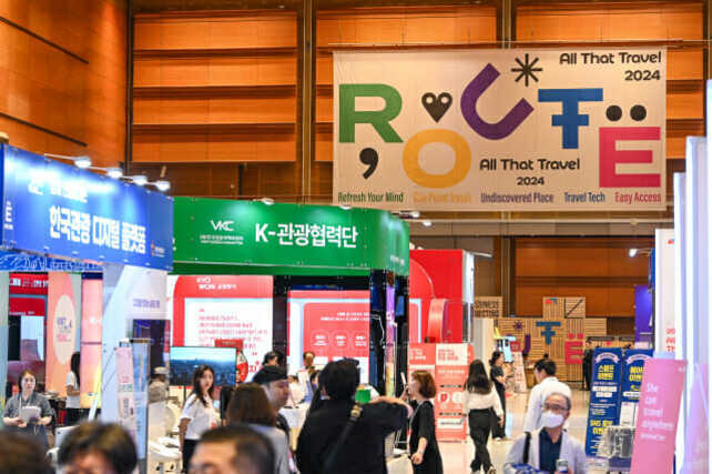 여러 사람들이 참석한 실내 여행 박람회 현장으로, 다양한 부스와 'ROUTE 2024 All That Travel'이라는 대형 배너가 보인다.