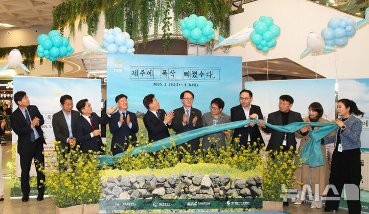 여러 명의 사람들이 제주도 홍보 행사에서 파란 천을 잡고 기념 촬영을 하고 있다. 뒤에는 유채꽃과 돌담이 있는 배경, 하늘색 풍선 장식 그리고 '제주에 푹 빠졌습니다'라는 문구가 적힌 현수막이 걸려 있다.
