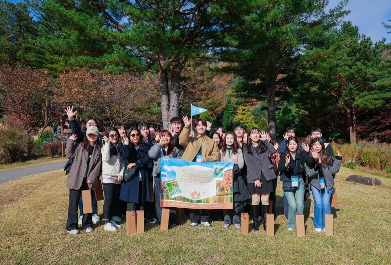 야외 공원에서 여러 명의 젊은 남녀가 모여 단체 사진을 찍고 있다. 가운데에는 '이화동의 아름다운 교실'이라고 쓰인 현수막을 들고 있으며, 모두 밝게 손을 흔들며 미소 짓고 있다. 주변엔 푸른 소나무와 가을 풍경이 펼쳐져 있다.