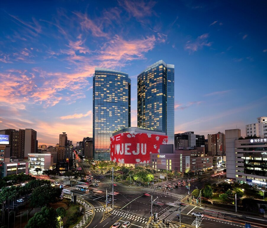 도심의 교차로와 고층 빌딩 두 개가 저녁 하늘 아래에 서 있고, 중앙 건물 외벽에는 'WE JEJU'라는 문구가 대형 전광판에 표시되어 있다.
