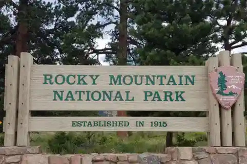 록키 마운틴 국립공원(Rocky Mountain National Park)이라고 적힌 나무 표지판이 숲을 배경으로 세워져 있다. 표지판에는 1915년에 설립되었다는 문구도 보인다.