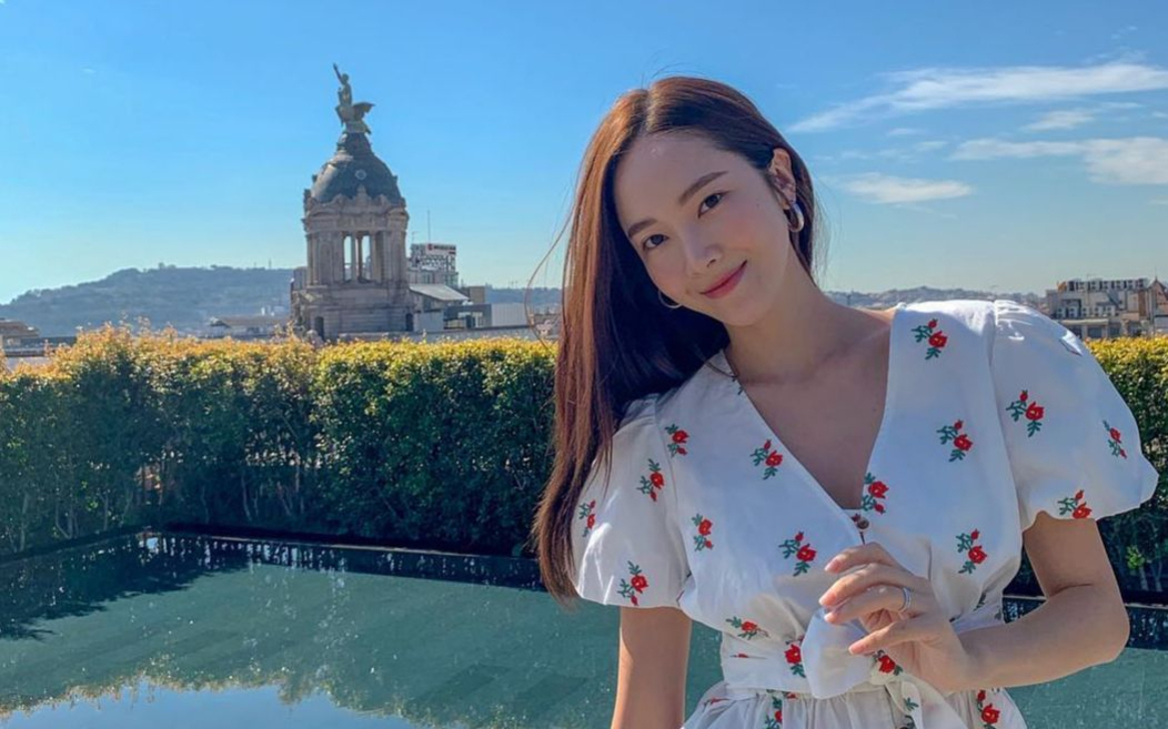 흰색 꽃무늬 원피스를 입은 여성이 야외에서 미소 지으며 포즈를 취하고 있다. 뒤에는 푸른 하늘과 고풍스러운 건물, 그리고 나무 울타리가 보인다.