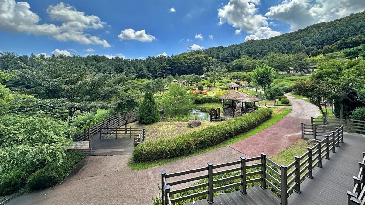 로컬 여행지 숨은 명소 추천: 서울 근교부터 지방 소도시까지 특별한 자연명소 탐방법 2 푸른 산과 숲으로 둘러싸인 공원 풍경. 산책로와 나무 울타리, 연못, 정자와 물레방아가 보인다. 맑은 하늘에 구름이 떠 있다.
