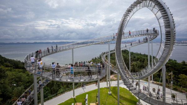 사람들이 걷고 있는 독특한 루프 모양의 전망대 산책로가 바다와 숲을 배경으로 설치되어 있다.