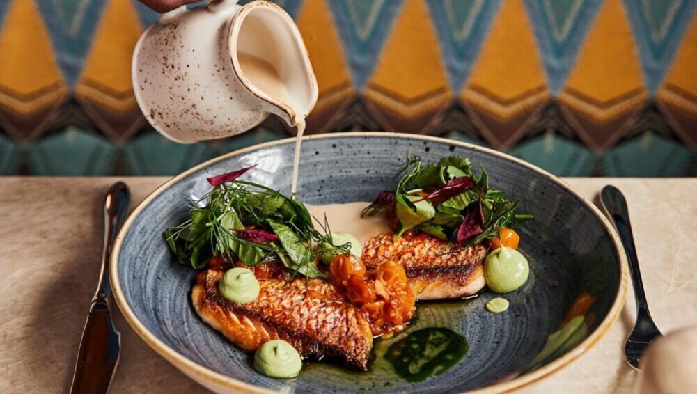 푸른 접시에 구운 생선 필레와 다양한 채소, 초록색 소스가 담겨 있고, 작은 주전자에서 크림 소스를 붓는 장면.