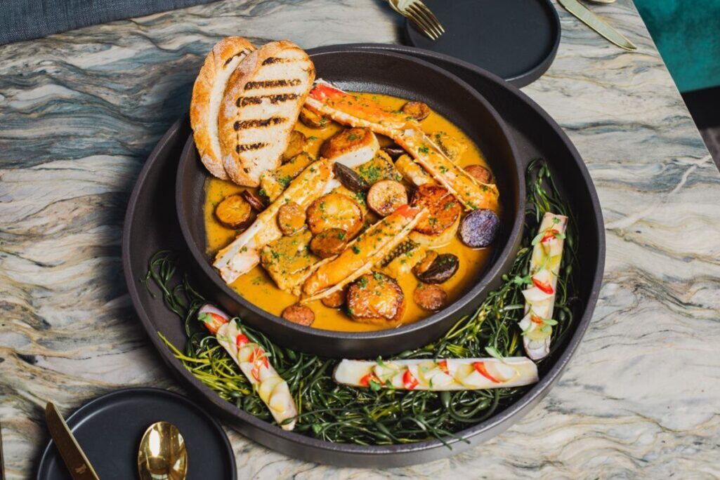 구운 빵 두 조각과 감자, 게 다리 등이 담긴 크림 소스 요리가 큰 접시에 플레이팅되어 있고, 주변에는 해초와 롤 형태의 음식이 장식된 고급스럽고 예술적인 한 끼 식사.