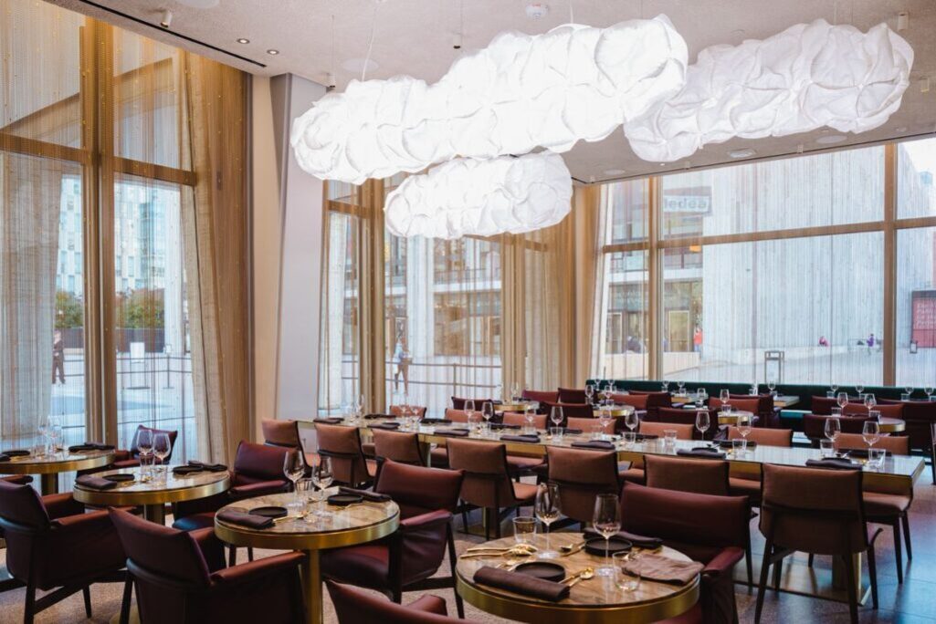 고급스러운 인테리어의 레스토랑 내부, 천장에는 구름 모양 조명이 달려 있고 테이블마다 식기와 와인잔이 세팅되어 있다. 큰 창문을 통해 자연광이 들어오며 외부 풍경도 보인다.