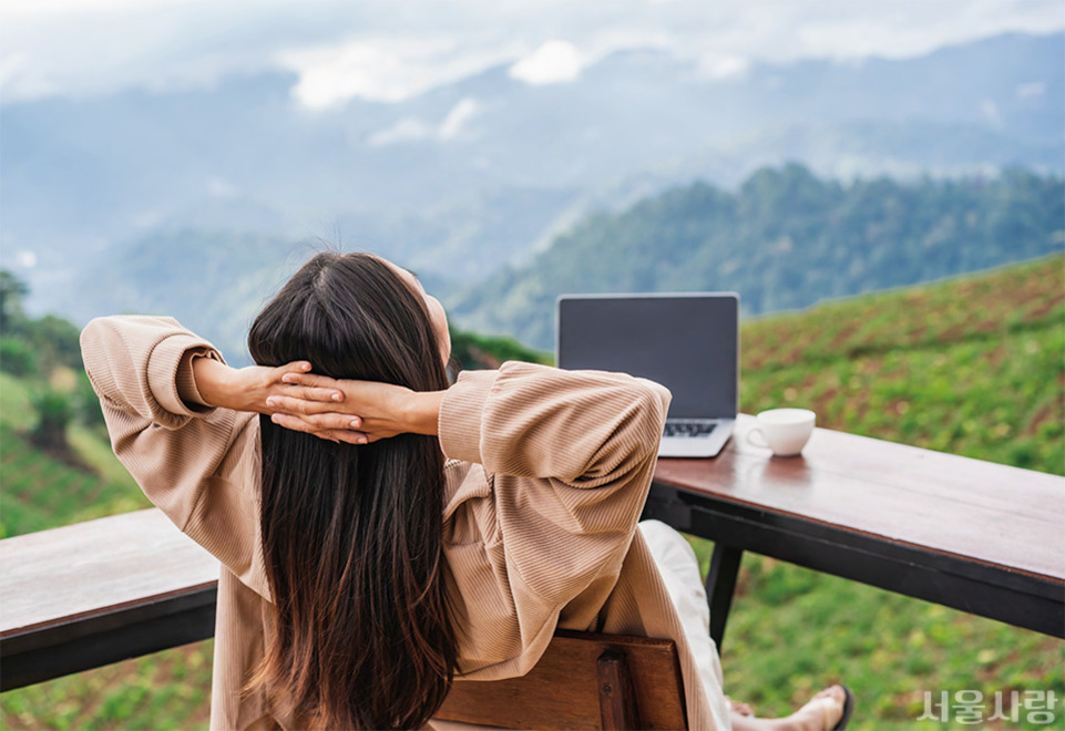 여성이 야외 테이블에 앉아 두 손을 머리 뒤로 하고 경치를 감상하고 있다. 책상 위에는 노트북과 커피잔이 놓여 있으며, 배경으로는 푸른 산과 들판이 보인다.